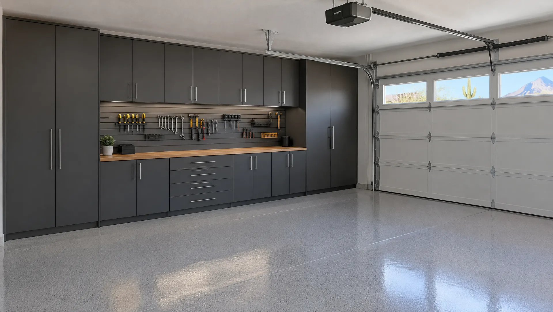 Phoenix home garage with wall-to-wall charcoal gray garage cabinets, a wood workbench top, slatwall panel with hanging tools, epoxy floor in light gray, a clean two-car garage with the door closed